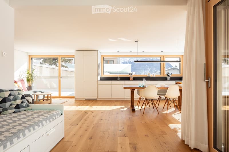 Open-plan living and dining area with light wood flooring, white walls, and large windows overlooking a snowy mountain view. Features a sofa, dining table with chairs, kitchen area with cabinets and a sink, and a potted plant near the window.