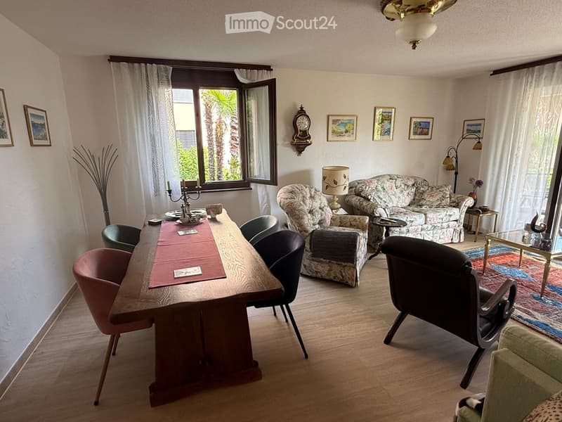 A well-furnished living room with a large wooden dining table, multiple chairs, a patterned sofa, a side chair, a floor lamp, a wall clock, and several picture frames on the wall. The room has large windows with white curtains and a view of palm trees out