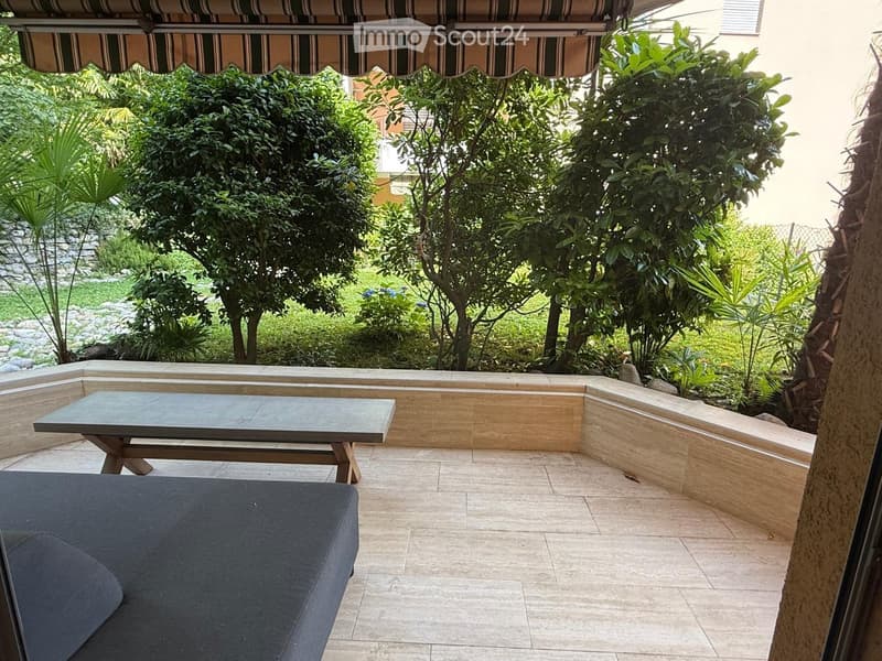 A well-maintained outdoor patio with a stone seating area, a wooden table, and a dark-colored sofa, surrounded by lush greenery and trees.