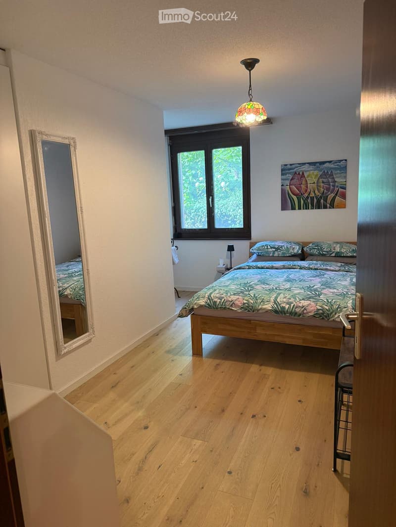 Cozy bedroom with wooden flooring, a double bed with a colorful duvet, a mirror, a small table lamp, and a painting on the wall.