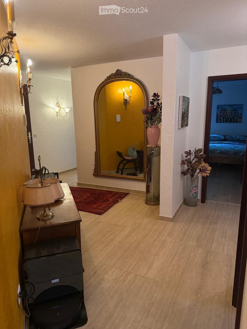 Elegant hallway with wooden flooring, a large ornate mirror, a table with a lamp, and vases with flowers. A doorway leads to a bedroom.