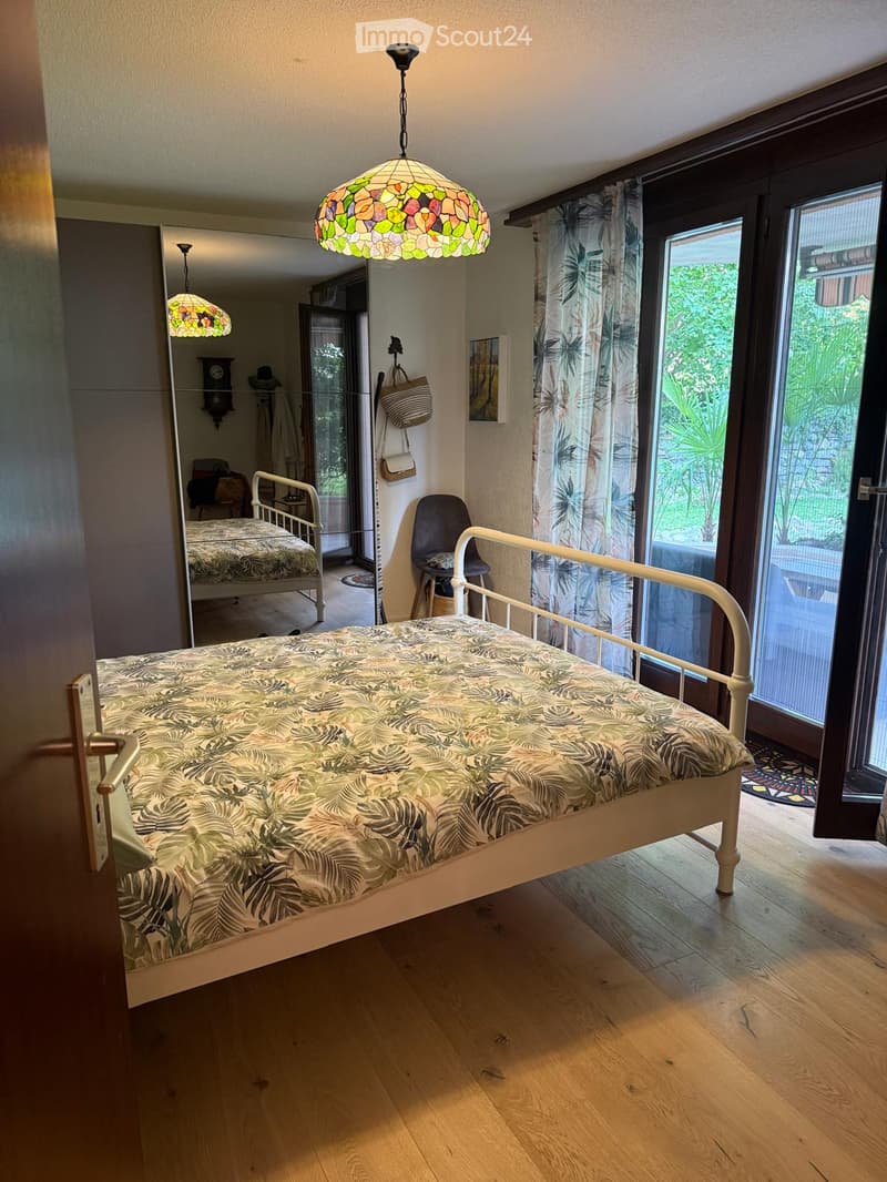 Bright bedroom with wooden flooring, a single bed with a leafy-patterned duvet, a hanging stained-glass lamp, a chair, a mirror, and a view of greenery through large windows.