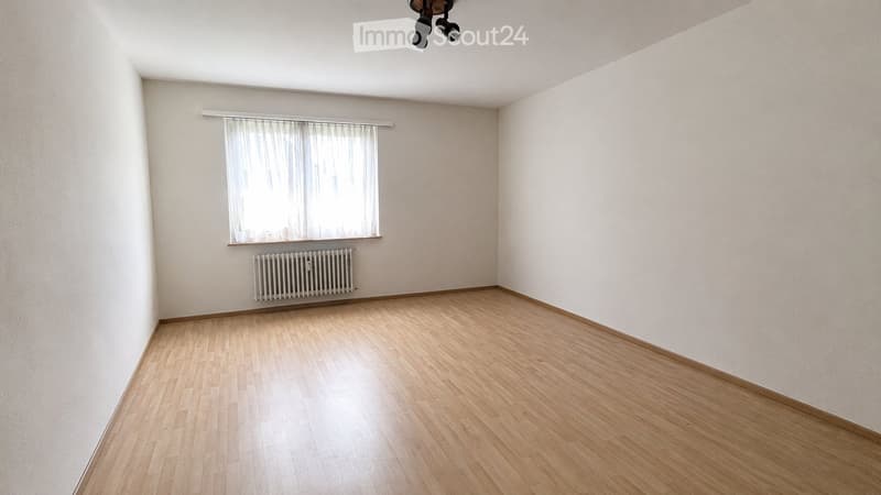 Empty room with wooden flooring, white walls, a window with white curtains, and a radiator below the window.