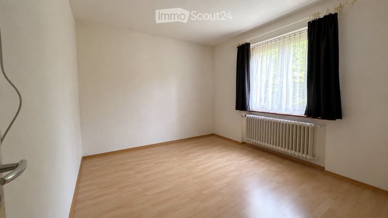 Empty room with wooden flooring, white walls, and a window with black curtains and a radiator below.
