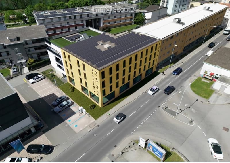 Aerial view of a modern multi-story residential building with solar panels on the roof, surrounded by parked cars and greenery.