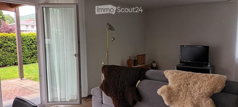 Modern living room with grey sofa, throw blankets, television on a stand, floor lamp, and decorative items on a small table. Large window with white curtains offers a view of a green garden.