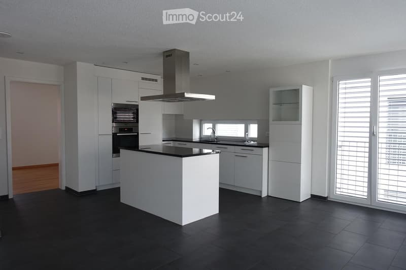 Modern kitchen with white cabinets, black countertops, a central island, and built-in appliances including an oven and range hood. Large windows with blinds provide natural light.