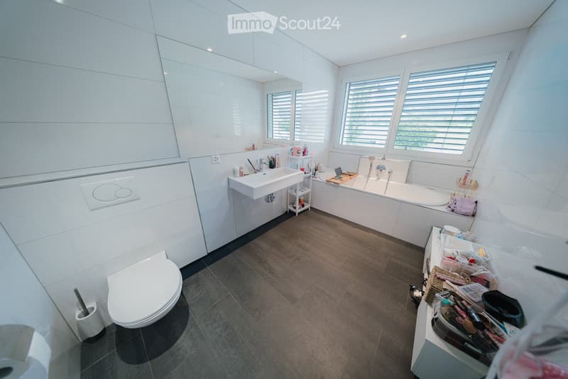 Modern bathroom with white tiles, a toilet, sink, bathtub, and a large mirror. The space includes storage cabinets and a shower rack with toiletries.