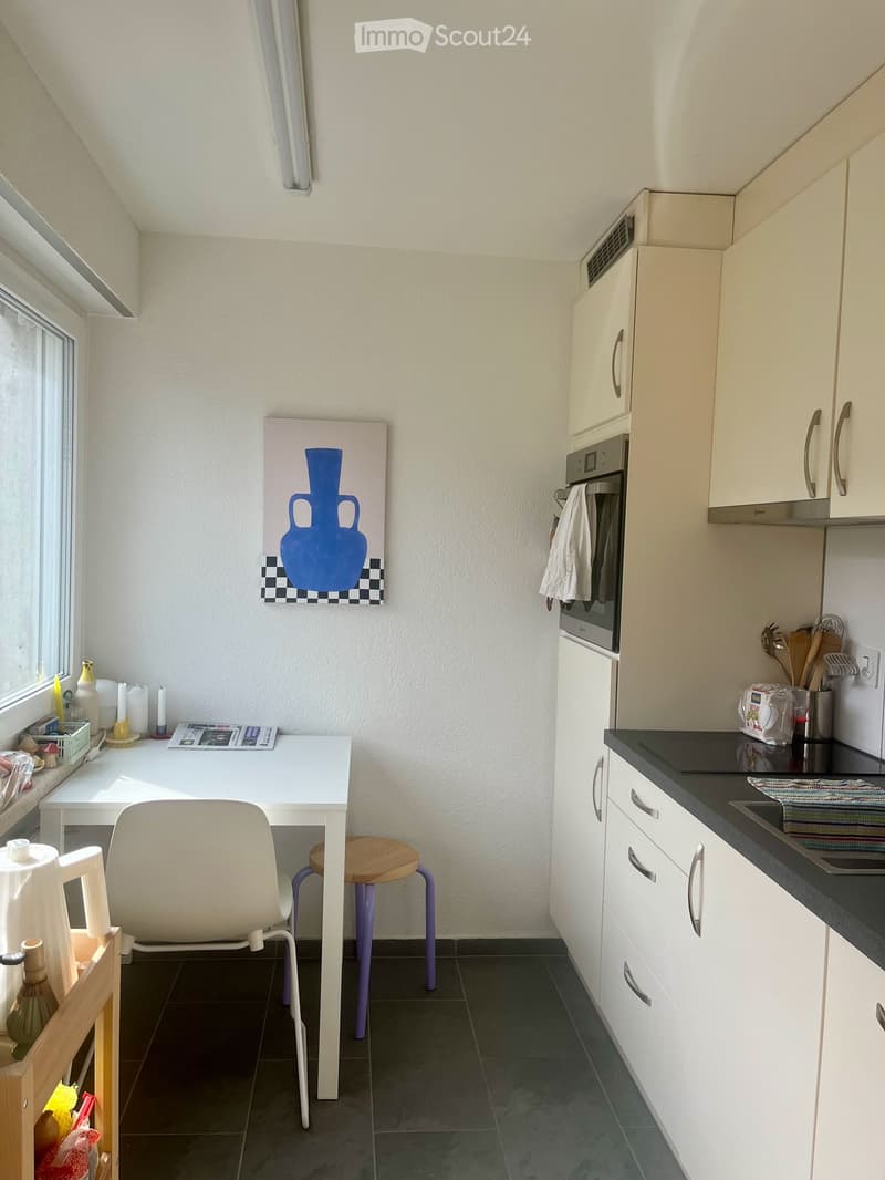 A compact kitchen with white cabinets, a black countertop, a sink, and an oven. A small white table with a chair and a stool is positioned near a window, with a painting on the wall and various small items on the table and counter.
