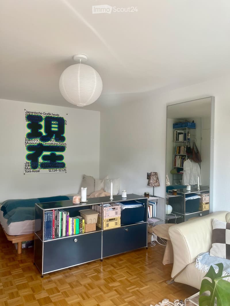 Modern furnished room with wooden flooring, a sofa, a bed, a cabinet, a small table, a lamp, and a mirror. The room features a large poster on the wall and a hanging spherical light fixture.