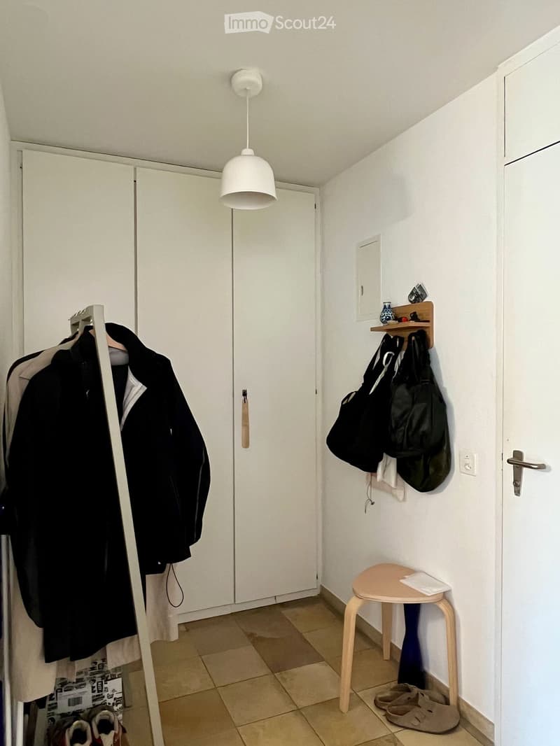 A narrow hallway with white walls, tiled floor, and a hanging pendant light. Features include a coat rack with jackets, a small wooden stool with a folded towel, and a shelf with bags and shoes.