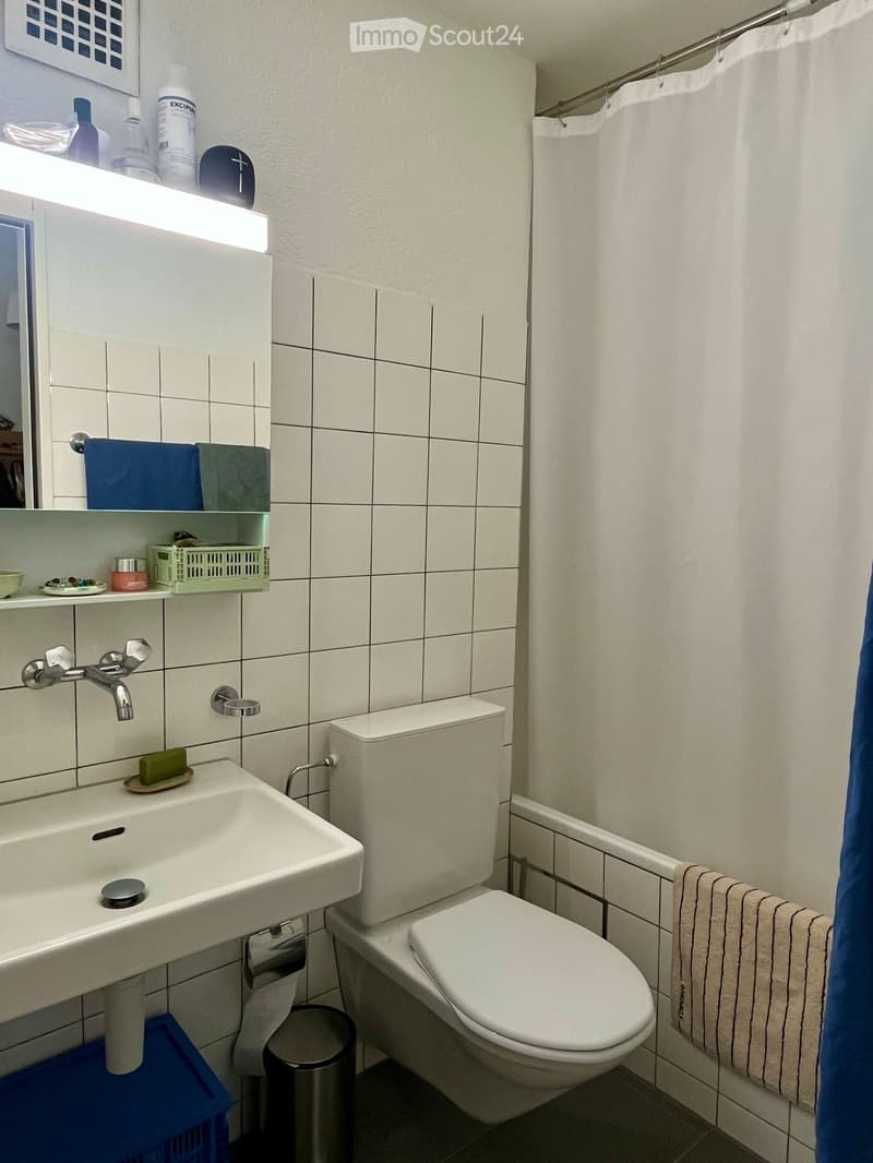 Modern bathroom with white tiles, a sink, toilet, mirror, and shower curtain. Shelves hold various toiletries and a towel is draped on the side.