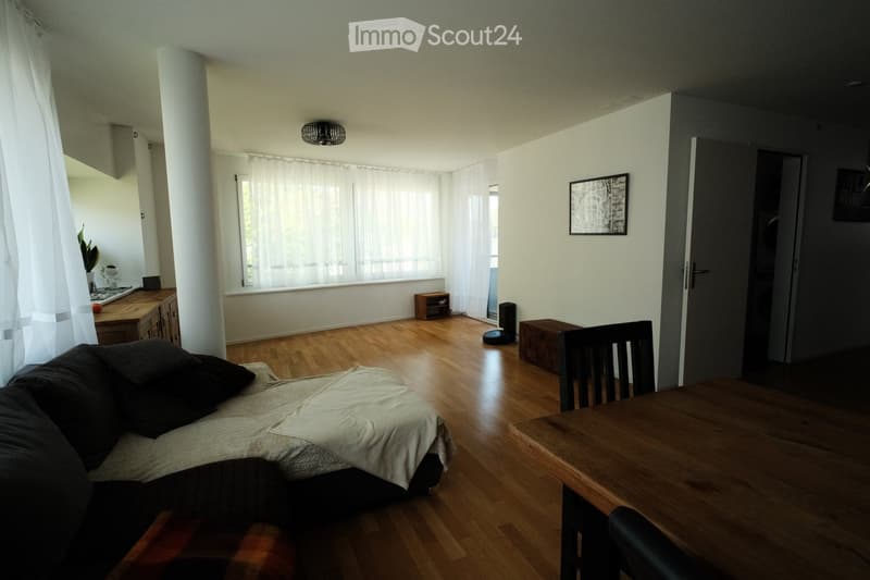 A well-lit living room with wooden flooring, a sofa, a dining table with chairs, a cabinet, a lamp, and a plant near the window.