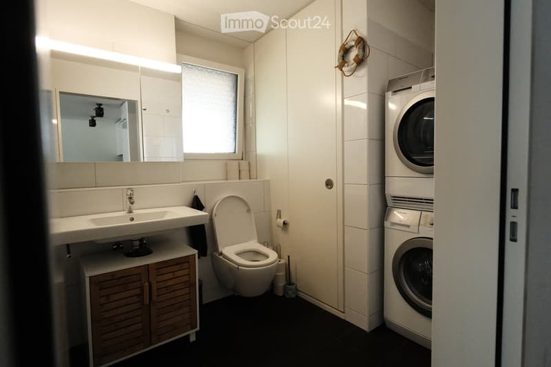Modern bathroom with a sink, mirror, toilet, and a washing machine. Features wooden cabinets, a frosted window, and a dark tiled floor.