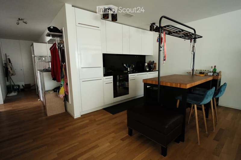 Modern kitchen with white cabinets, a wooden island, and a black oven. The space includes a bar table with two chairs, a stool, and a hanging rack with various items.