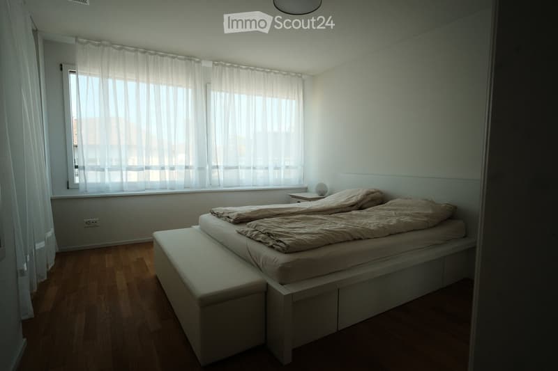Minimalist bedroom with a white bed, beige bedding, wooden floor, and large window with sheer curtains.