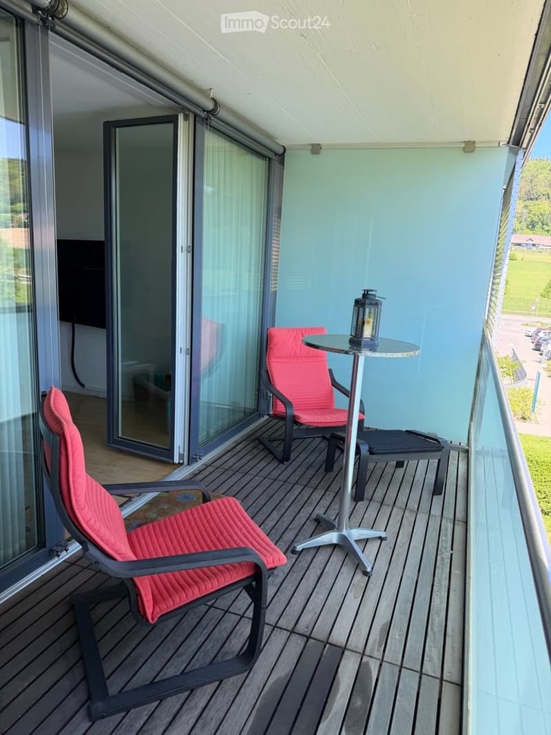 Balcony with wooden decking, two red chairs, a small round table, and a lantern. Large glass sliding doors open to the interior.