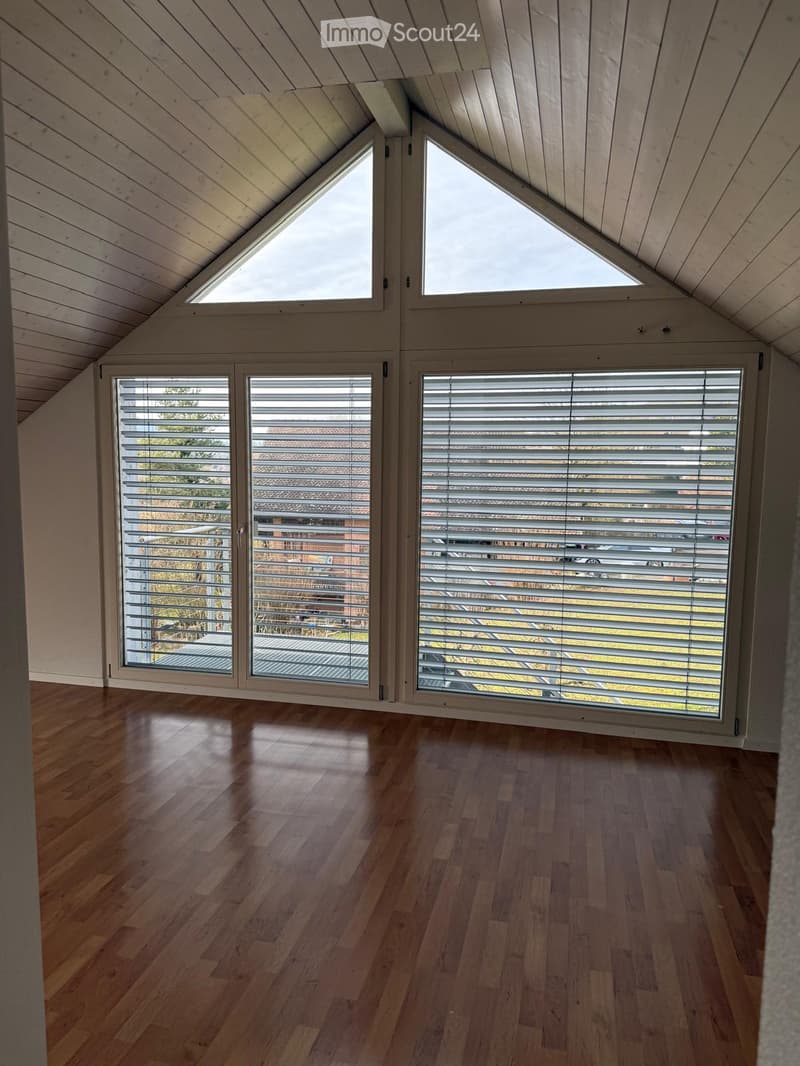 Bright, airy attic room with wooden floors, large windows, and skylights, featuring a sloped wooden ceiling and modern blinds.