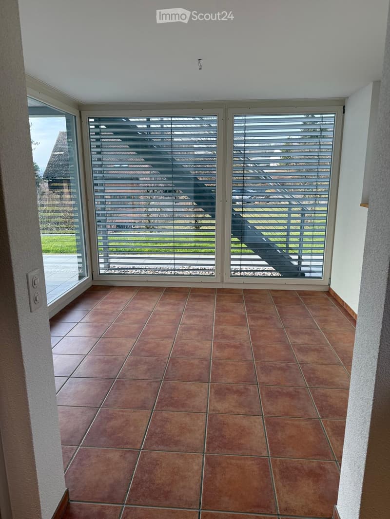 Empty room with brown tiled floor and large windows with blinds, leading to an outdoor area.