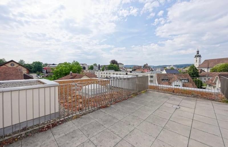 Rooftop terrace with metal railing, tiled floor, and scenic city view with buildings and a church tower.