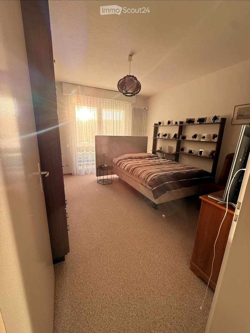 A well-lit bedroom with a bed, nightstand, shelf, chair, and a small table with a lamp. The room features carpeted flooring and large windows with sheer curtains.