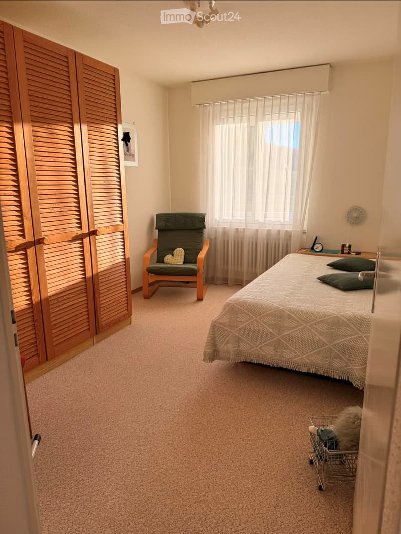 Cozy bedroom with a double bed, green pillows, a wooden wardrobe, a green armchair with a heart-shaped cushion, and a small nightstand with a clock.
