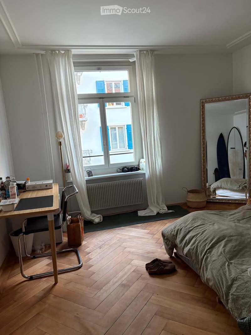 Cozy bedroom with wooden flooring, large window with white curtains, bed with green bedding, desk with chair, mirror, and surfboard in the corner.