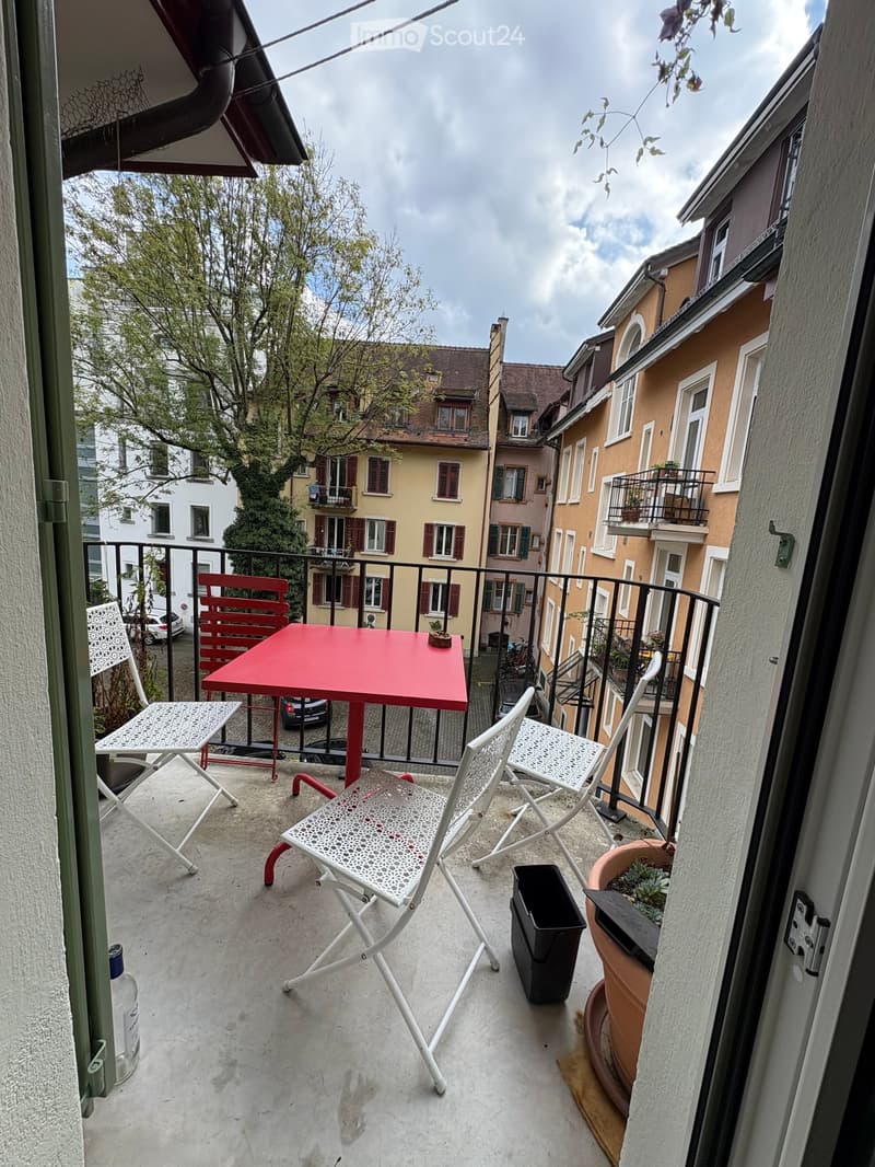 Balcony with a red table, four white chairs, a potted plant, and a trash bin. The balcony overlooks a street with buildings and trees.