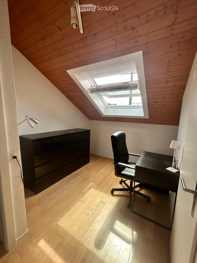 Modern, well-lit room with wooden flooring, a black desk, a black office chair, a black cabinet, and a small lamp on the desk. A skylight provides natural light.