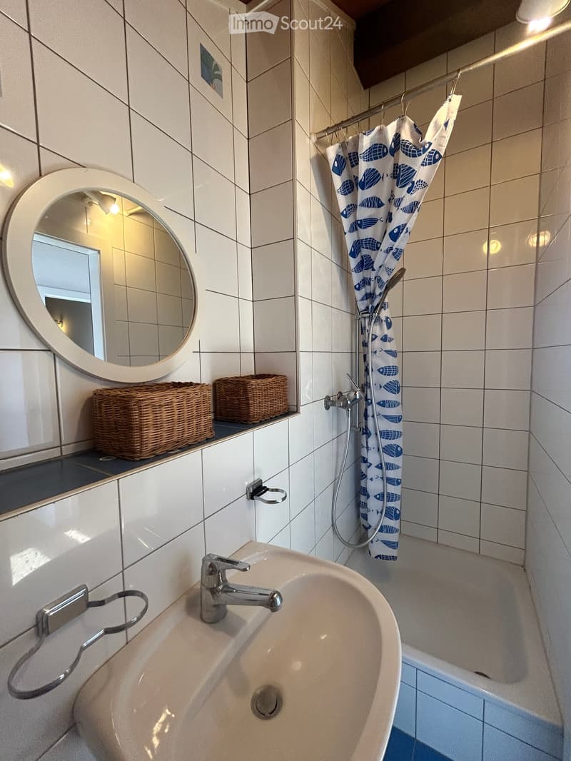 Bathroom with white tiled walls, a sink with a modern faucet, a bathtub with a blue fish-patterned shower curtain, a round mirror, and two wicker baskets on a shelf.