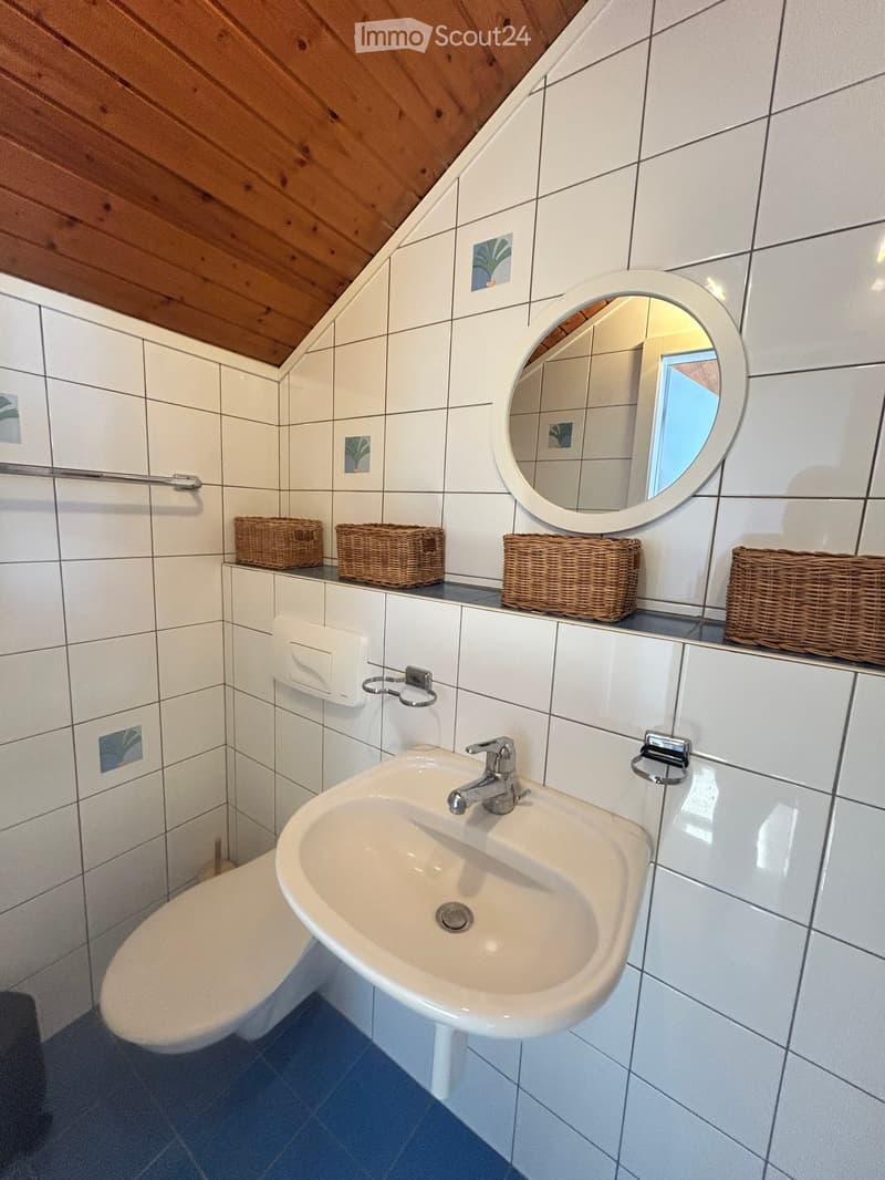 Bathroom with white tiled walls, a sink with a chrome faucet, a toilet, a round mirror, and wicker baskets on the counter.