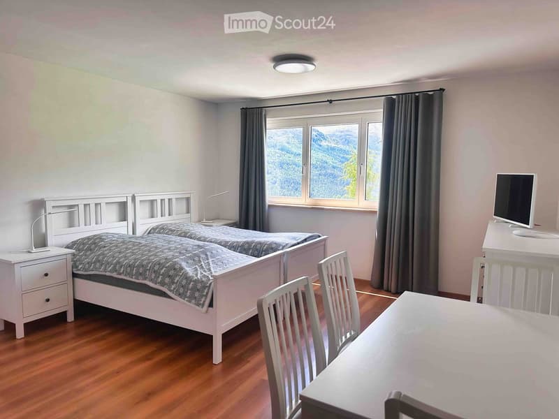 Cozy bedroom with two single beds, nightstands, a desk with a computer, and chairs near a large window with a scenic mountain view.