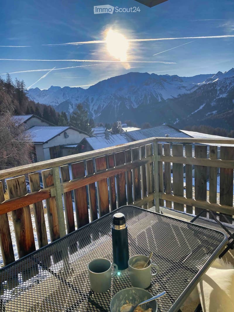Balcony with wooden railing, metal table, cups, and a bowl, overlooking snow-capped mountains and a village.