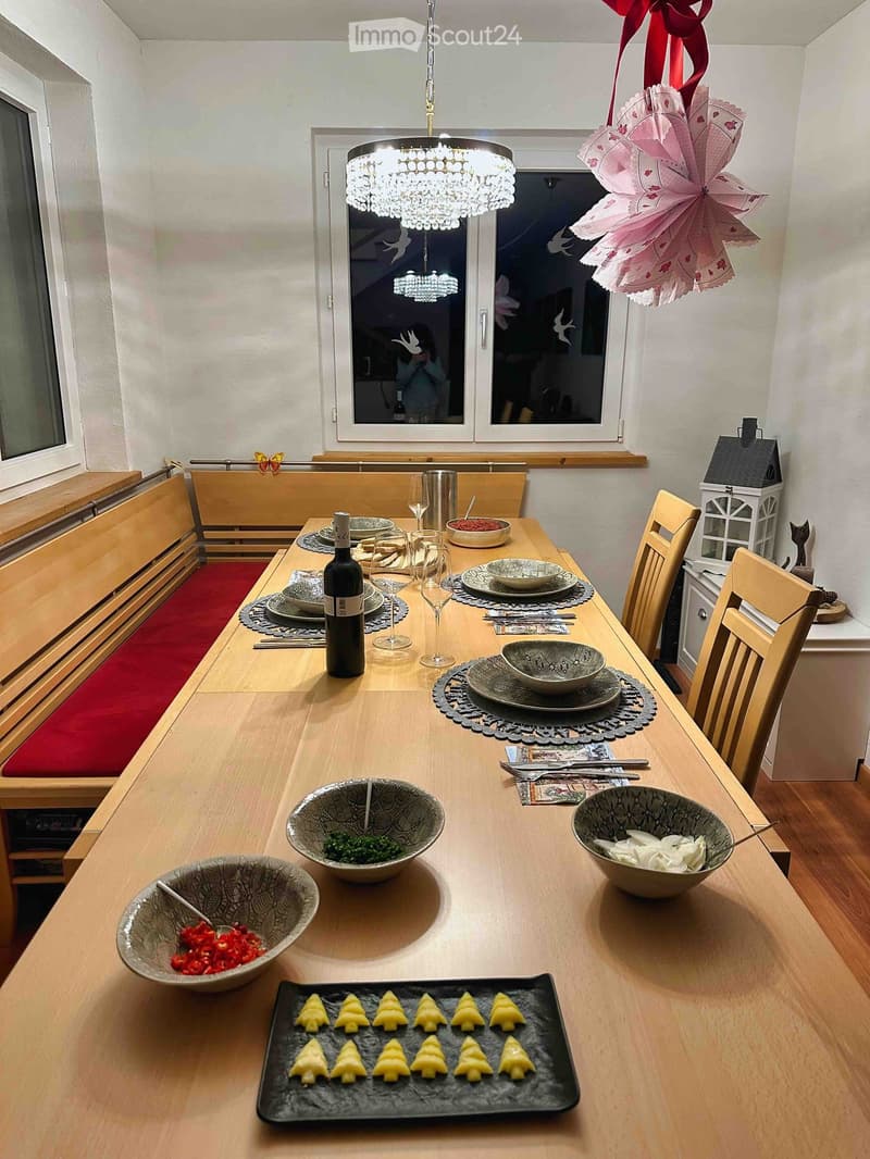 A wooden dining table set with plates, bowls, cutlery, and a bottle of wine, surrounded by wooden chairs and a chandelier hanging from the ceiling.