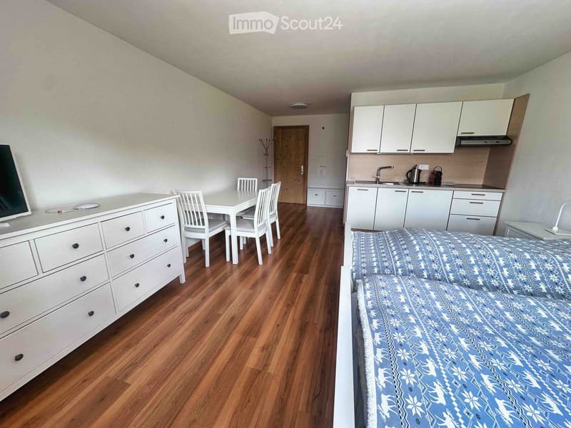 A furnished room with a bed, dresser, small dining area with table and chairs, and a compact kitchen area with cabinets and sink.