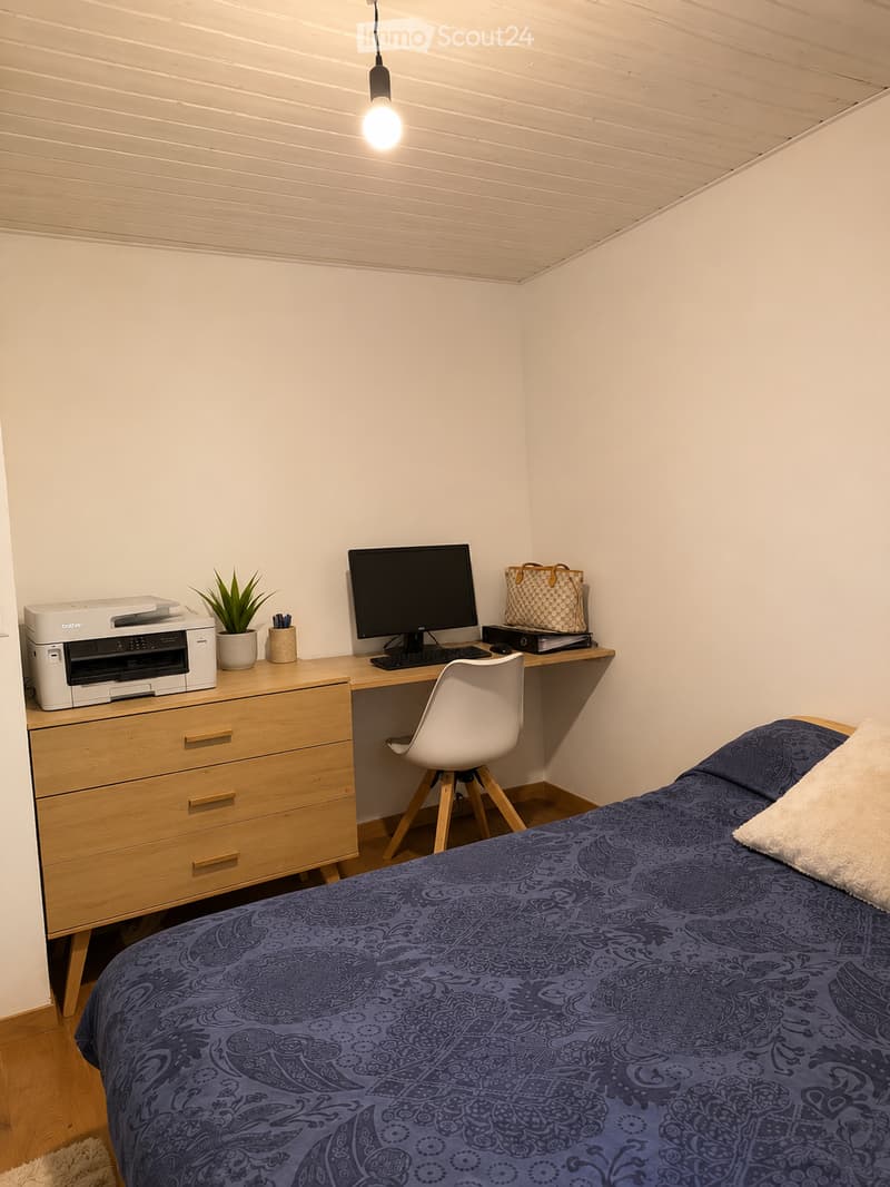 Cozy bedroom with a double bed, wooden desk, white chair, printer, and small plant on the desk.
