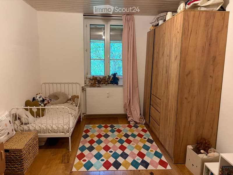 Cozy children's bedroom with a white metal bed, colorful rug, wooden wardrobe, and stuffed toys on the windowsill and bed.