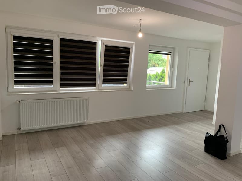 Empty room with light-colored walls, wooden flooring, and large windows with blinds. A radiator is visible on one wall, and a black bag is placed near the corner.