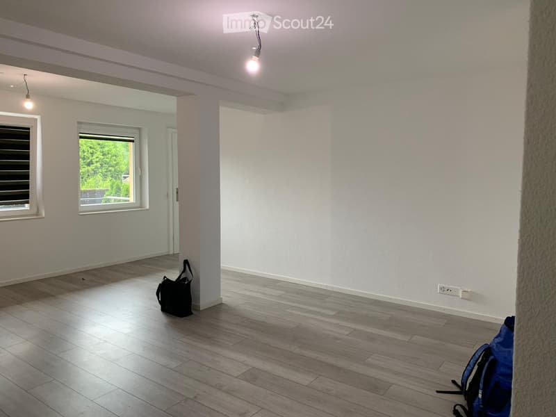 Empty room with light wooden flooring, white walls, and two windows. A black handbag and a blue backpack are placed on the floor.