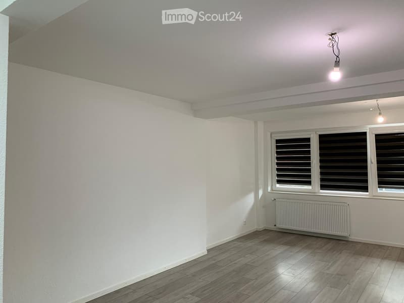 Empty room with white walls, wooden flooring, and large windows with blinds. Features a radiator and ceiling lights.