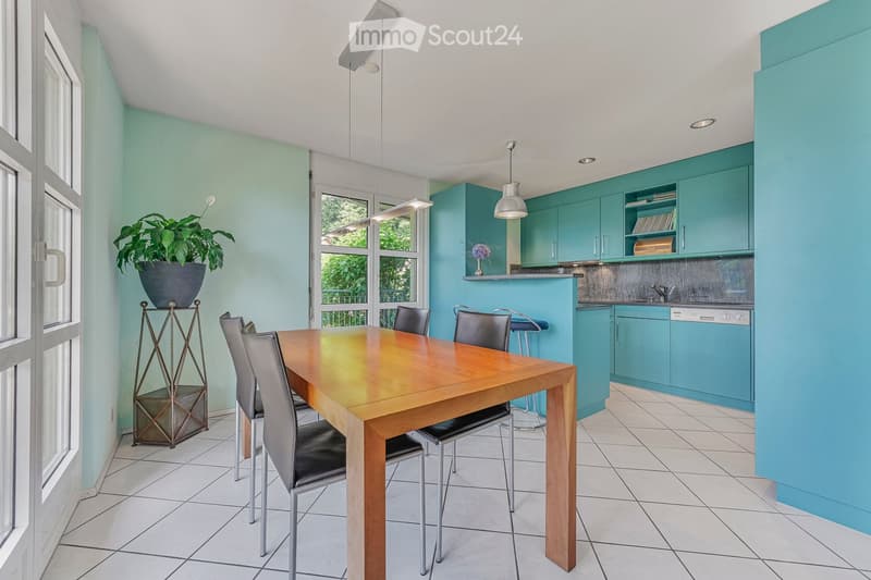 Bright dining area with a wooden table, six chairs, and a stool near the kitchen. The kitchen features turquoise cabinets, a dishwasher, and a countertop with a vase and plant. The room has large windows and modern lighting.