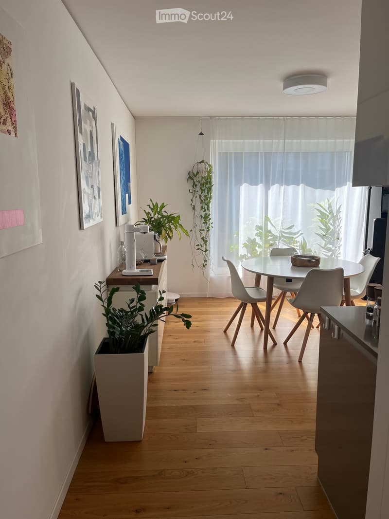 A modern, well-lit room with wooden flooring, a round dining table with four chairs, a coffee machine on a small counter, and several potted plants. The room features large windows with sheer curtains and artwork on the walls.