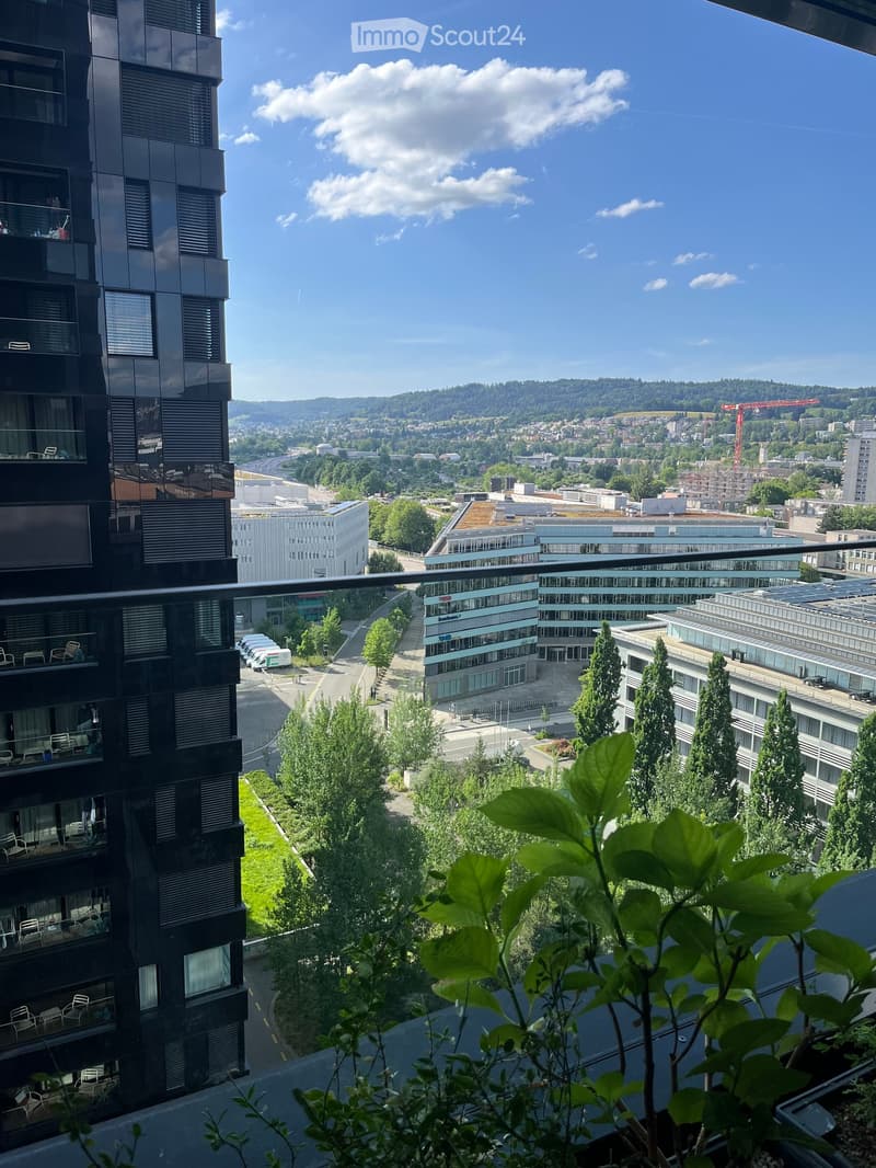 Balcony overlooking a cityscape with modern buildings, greenery, and a clear blue sky. A few chairs are visible on the balcony.