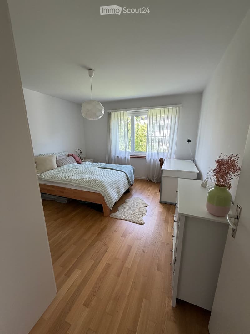 Bright bedroom with wooden flooring, a bed with striped bedding, a nightstand with a lamp, a small table with a vase, and a white rug on the floor.