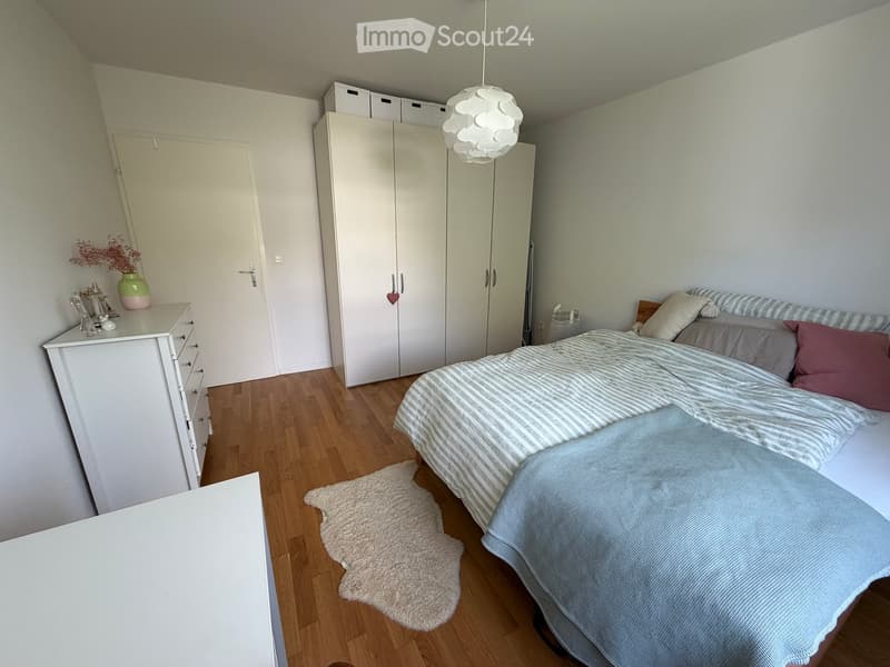 Cozy bedroom with wooden flooring, a bed with striped bedding, a white wardrobe, a nightstand with decorative items, and a small rug.