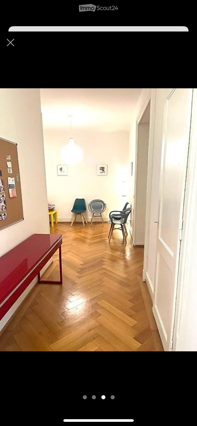 A narrow hallway with wooden herringbone flooring, white walls, and modern lighting. Contains a red bench, a yellow stool, and several chairs.