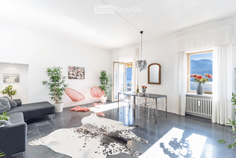 Bright, modern living room with white walls, large windows offering a scenic view, grey sofa, coffee table, pink chairs, glass dining table, and decorative plants.