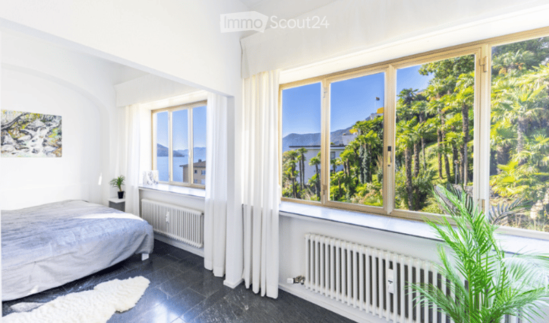 Bright bedroom with large windows offering a scenic view, featuring a bed, radiator, curtain, small plant, and a picture frame on the wall.