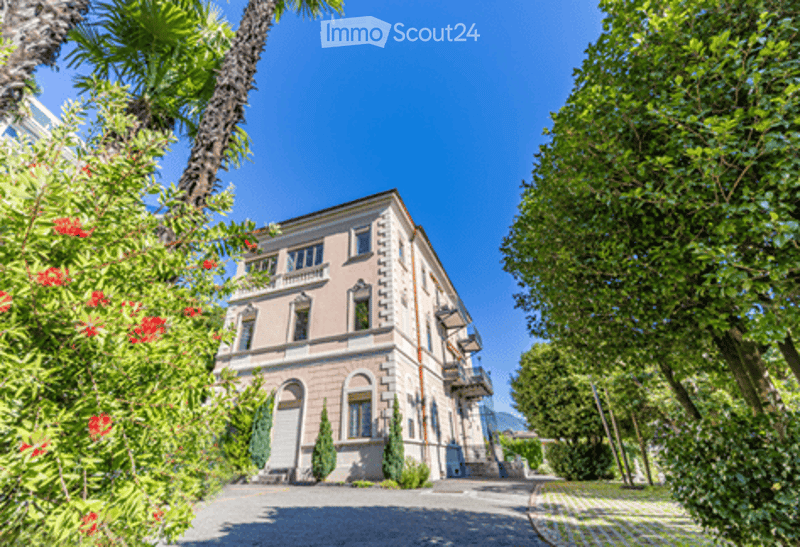 A large, elegant villa with classical architecture, surrounded by lush greenery and palm trees. The building features arched windows, a symmetrical facade, and a well-maintained garden.