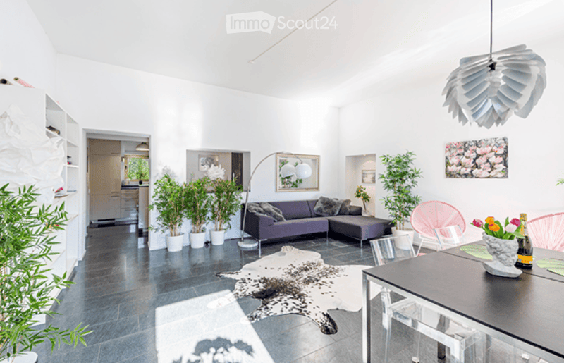 Modern living room with white walls, dark grey sofa, coffee table, dining table, chairs, floor lamp, artwork, and plants.